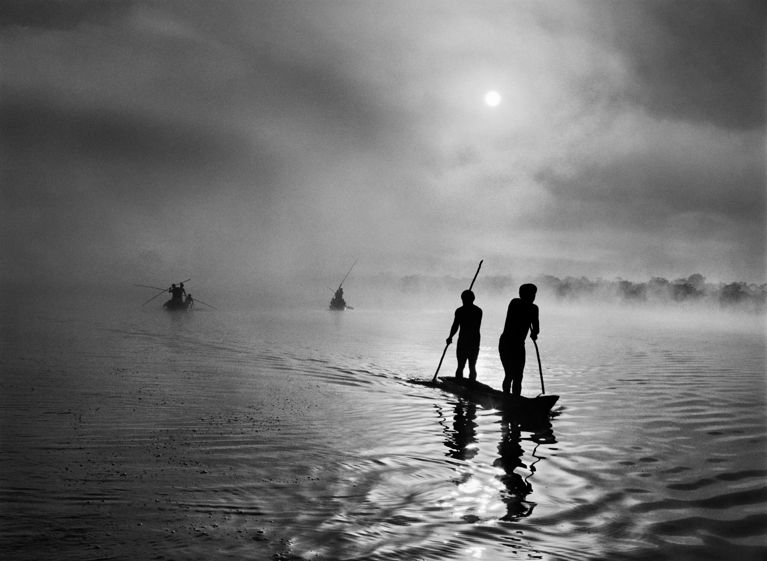 Eine Schwarz-Weiß-Fotografie des Fotografen Sebastião Salgado zeigt mehrere Personen, die auf einem Fluss paddeln.