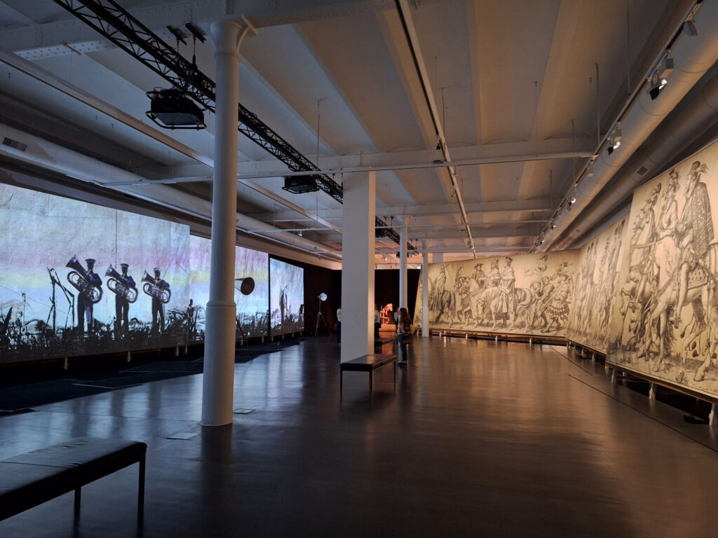 Zu sehen ist ein Ausstellungsraum mit einer Videoinstallation von William Kentridge links und den großformatigen Kartons des Fürstenzugs rechts.