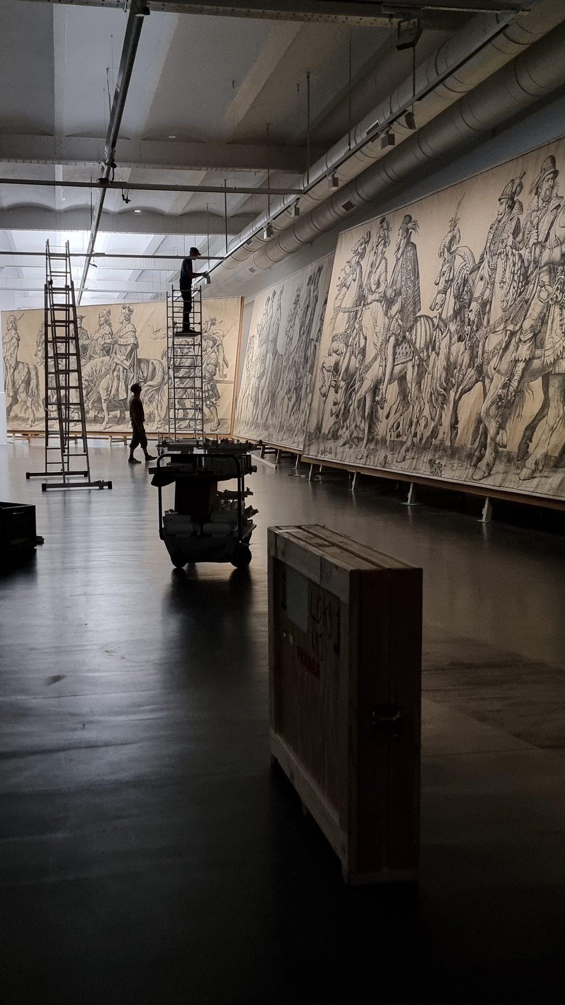 Zu sehen ist ein großer Ausstellungsraum, in dem sich zwei Personen befinden, eine steht auf einer Leiter vor den großformatigen Kartons.