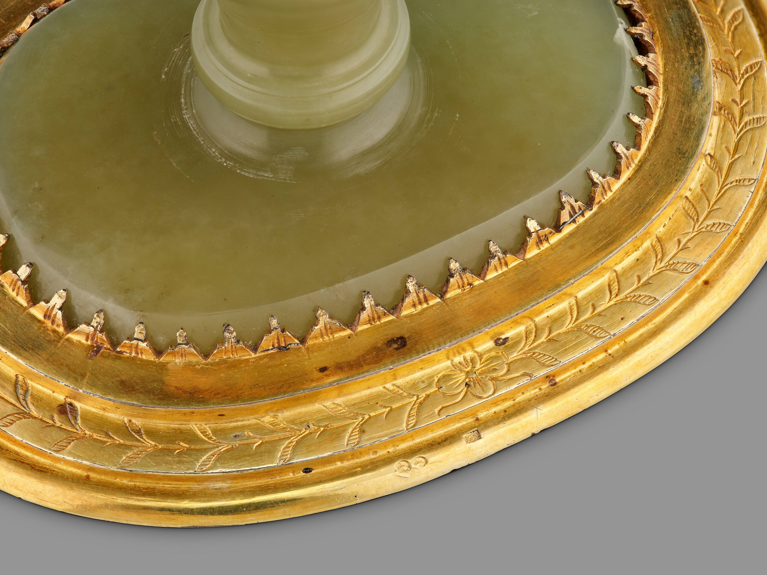 Zu sehen ist eine Detailansicht des Fußes der Kanne aus Nephrit mit silbervergoldeter Fassung mit Zackenrand, floraler Verzierung und Punzierung.